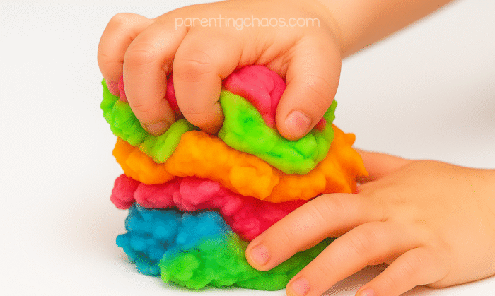 Child squishing a colorful stack of homemade playdough in bright neon colors — blue, pink, orange, green, and yellow — showing the soft, fluffy texture.