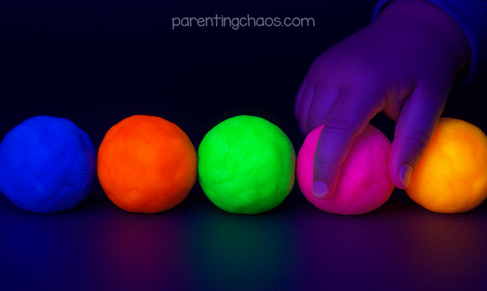 Child’s hand reaching for glowing playdough balls in blue, orange, green, pink, and yellow under black light.