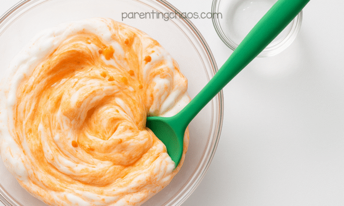 Green spoon stirring fluffy pumpkin pie slime ingredients together in the bowl.