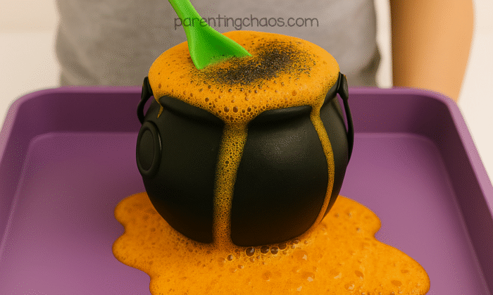 A child stirs an overflowing orange potion inside a black cauldron with a green spoon on a purple tray — part of a Halloween potion science experiment.