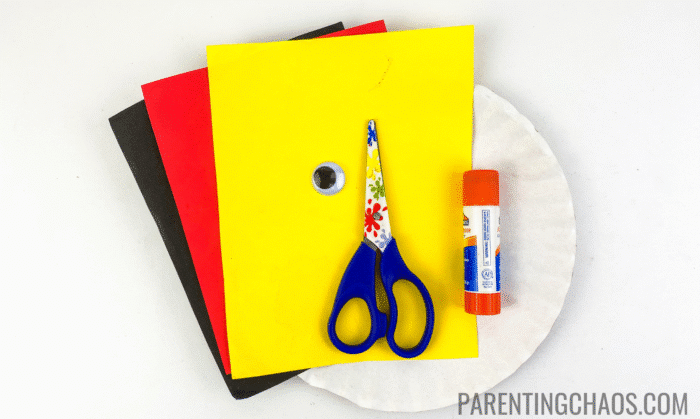 Colorful craft supplies for a Little Red Hen paper plate craft, including red, yellow, and black construction paper, a googly eye, child-safe scissors with a floral pattern, an orange glue stick, and a white paper plate, all arranged on a white background.
