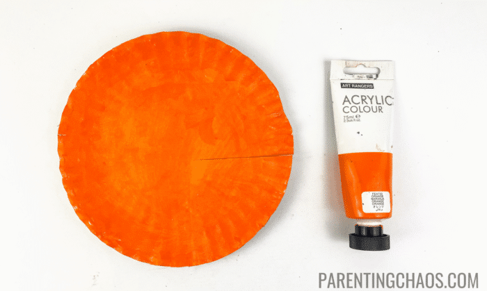 Orange-painted paper plate with a faint pencil line drawn to mark where to cut for the wing, shown next to a tube of orange acrylic paint on a white background for a Little Red Hen paper plate craft.