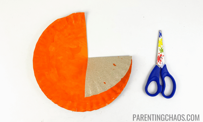 Orange-painted paper plate with a cut made along a marked pencil line and the folded section turned down to create a wing, shown next to a pair of blue child-safe scissors on a white background for a Little Red Hen paper plate craft.