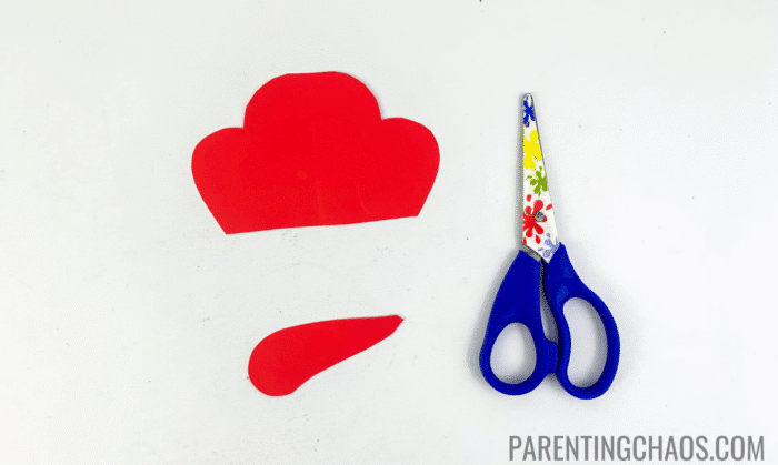 Red construction paper pieces cut into the Little Red Hen’s crest and beard shapes, placed next to a pair of blue child-safe scissors on a white background for a Little Red Hen paper plate craft.
