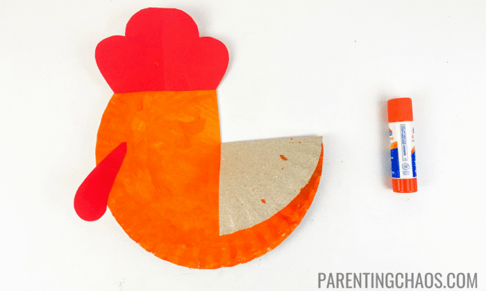 Orange-painted paper plate with red construction paper crest and beard glued in place, folded wing on the side, and an orange glue stick placed next to it, shown on a white background for a Little Red Hen paper plate craft.