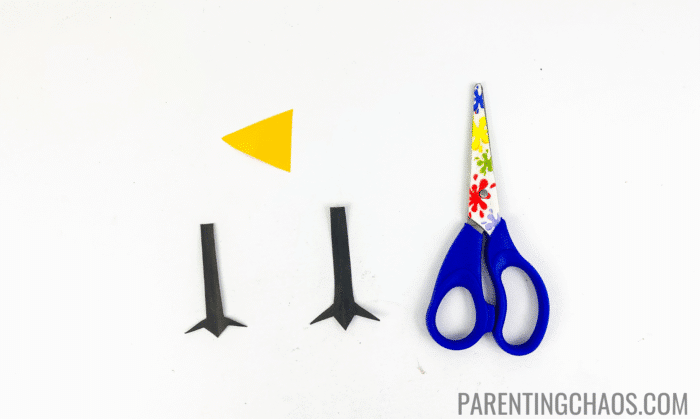 Small yellow triangle beak and two black cutout chicken feet made from construction paper, placed next to a pair of blue child-safe scissors on a white background for a Little Red Hen paper plate craft.
