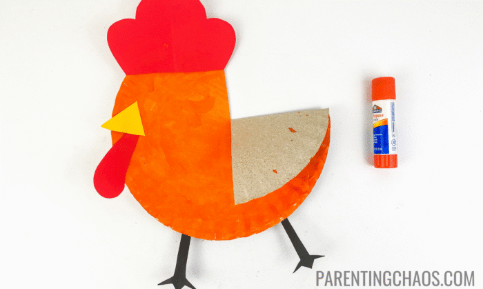 Nearly finished Little Red Hen paper plate craft with red crest and beard, yellow triangle beak, black paper feet, and folded wing, shown next to an orange glue stick on a white background.