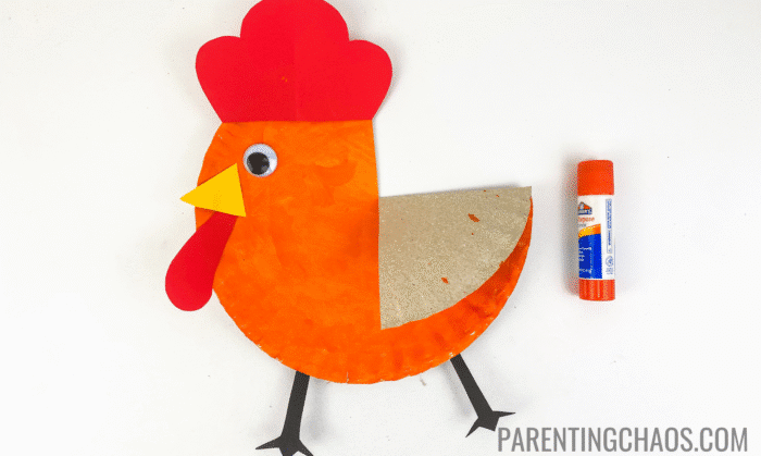 Completed Little Red Hen paper plate craft featuring an orange-painted plate, red crest and beard, yellow beak, googly eye, black paper feet, and folded wing, shown next to an orange glue stick on a white background.