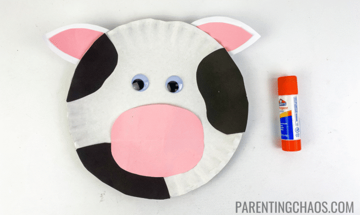 Cow paper plate craft with black spots, pink-and-white ears, a pink nose, and googly eyes, next to an orange Elmer’s glue stick.