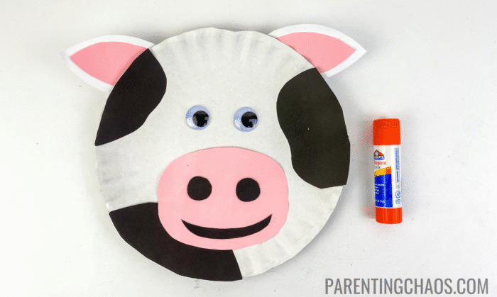 Finished cow paper plate craft with black spots, pink-and-white ears, pink nose with black nostrils, googly eyes, and a black paper smile, next to an orange Elmer’s glue stick.