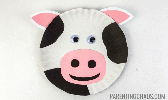Completed cow paper plate craft with black spots, pink-and-white ears, pink nose with black nostrils, googly eyes, and a black paper smile, displayed on a white background.