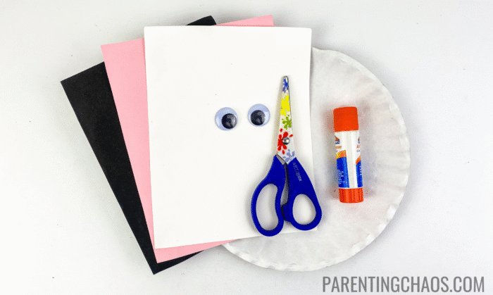 Supplies for a cow paper plate craft, including black, pink, and white construction paper, googly eyes, safety scissors, a glue stick, and a white paper plate, all laid out on a white background.