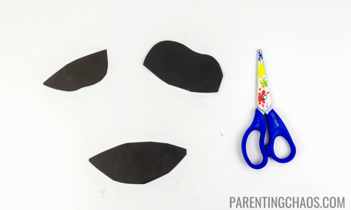 Black cow spots cut from construction paper next to blue safety scissors, ready to be used for a cow paper plate craft.