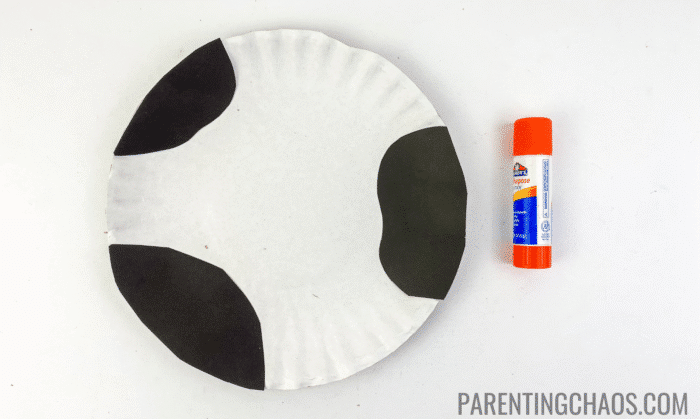 White paper plate with black construction paper cow spots glued on, next to an orange Elmer’s glue stick, part of a cow paper plate craft in progress.
