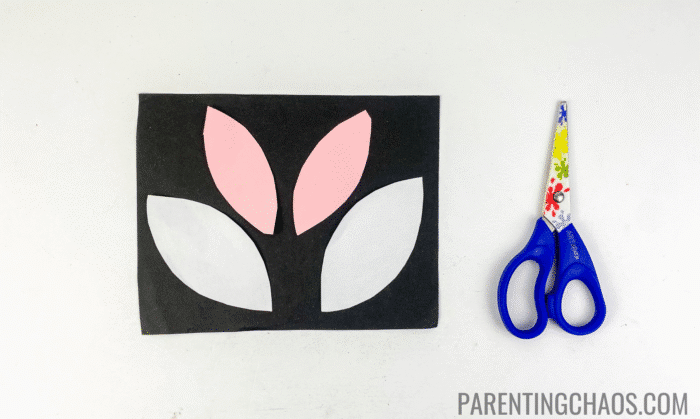 Pink and white paper cow ears cut from construction paper, placed on a black background, next to blue safety scissors for a cow paper plate craft project.