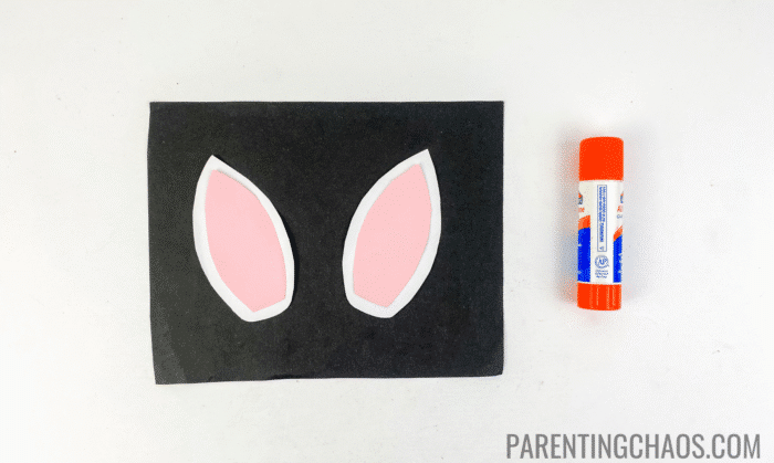 Two pink and white cow ears assembled from construction paper, placed on a black background next to an orange Elmer’s glue stick, part of a cow paper plate craft project.