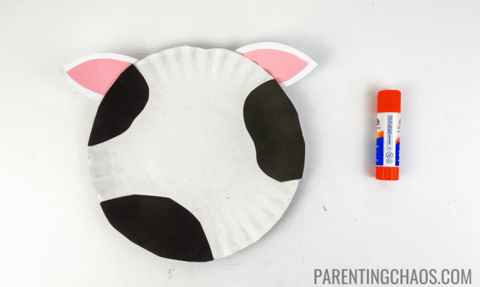 Cow paper plate craft in progress with black paper spots and pink-and-white paper ears glued onto a white paper plate, next to an orange Elmer’s glue stick.