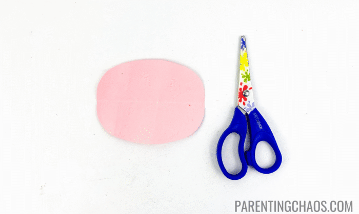 Pink construction paper cow nose cutout placed next to blue safety scissors, part of a cow paper plate craft project in progress.