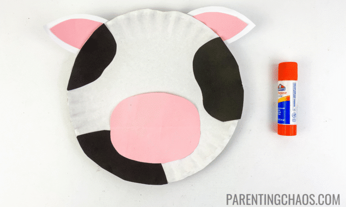 Cow paper plate craft with black spots, pink-and-white ears, and a pink nose glued onto a white paper plate, next to an orange Elmer’s glue stick.