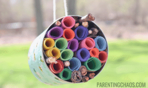 DIY bee hotel made from a recycled can filled with colorful rolled paper and sticks, hanging outside on a string with a green blurred background.