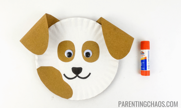 Completed paper plate dog craft with googly eyes, brown floppy ears, spots, and a smiling mouth, shown with an orange glue stick.