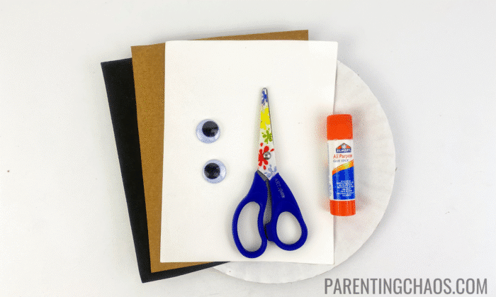 Supplies for a dog paper plate craft including scissors, glue stick, googly eyes, construction paper (brown, black, and white), and a plain paper plate.