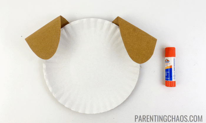 White paper plate with floppy brown paper ears glued to the top sides, shown with a glue stick for a dog paper plate craft.