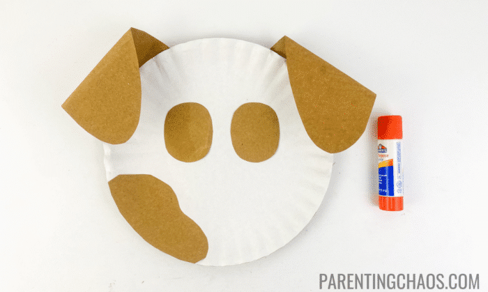 White paper plate with floppy brown ears, eye patches, and a large spot glued on as part of a dog face craft, shown with an orange glue stick.