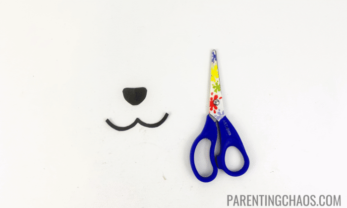 Black paper cutouts for a dog nose and mouth next to a pair of child-safe scissors, part of a dog paper plate craft activity.