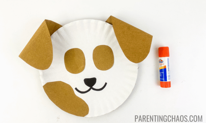 Paper plate dog craft with brown ears, eye patches, and a large spot, now showing the glued-on black nose and smiling mouth below, with a glue stick nearby.