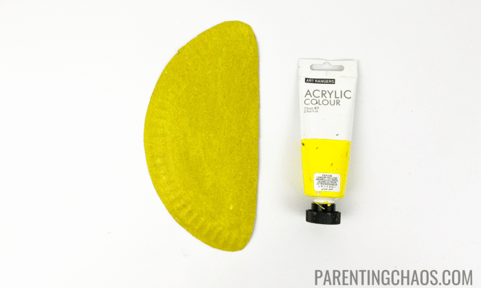 Half paper plate painted yellow next to a tube of yellow acrylic paint, part of a duck paper plate craft project.