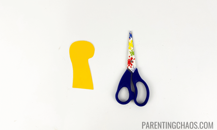 Yellow construction paper duck head cutout placed beside floral-patterned kids’ scissors, part of a paper plate duck craft.