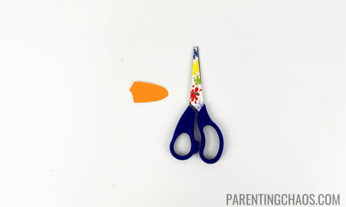 Orange duck beak cut from construction paper sitting next to colorful child-safe scissors, used in a preschool duck craft project.
