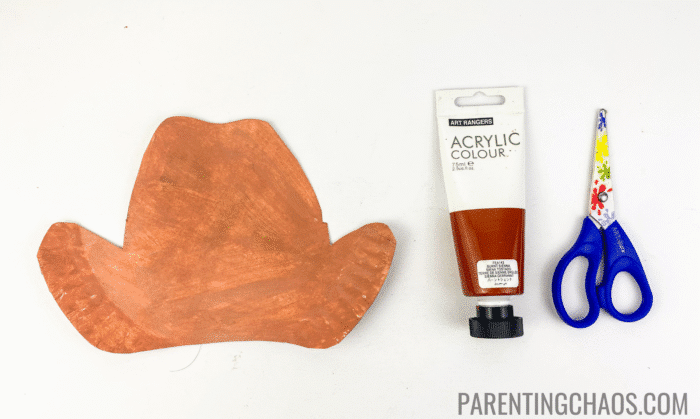 Partially completed paper plate cowboy hat craft painted brown, shown next to a tube of brown acrylic paint and blue kids’ scissors.