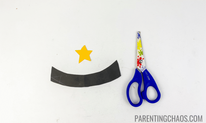 Cut-out black paper hatband and yellow sheriff star next to blue kids’ scissors, ready to be assembled for the paper plate cowboy hat craft.