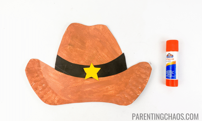 Finished paper plate cowboy hat craft painted brown with a black paper hatband and yellow sheriff star, shown next to an orange Elmer’s glue stick.