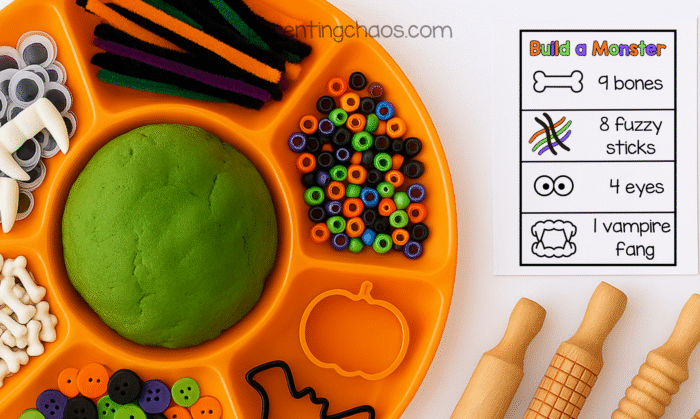 A Halloween playdough tray filled with green playdough, vampire fangs, googly eyes, bones, colorful beads, and pipe cleaners. A “Build a Monster” task card lists “9 bones, 8 fuzzy sticks, 4 eyes, 1 vampire fang.” Wooden rolling pins are on the side.