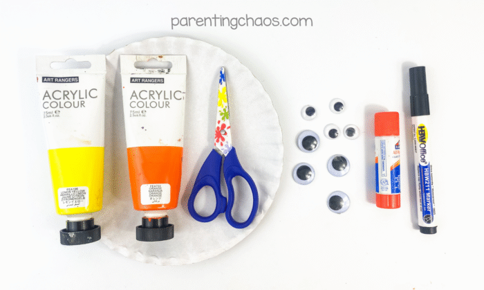 Supplies for candy corn paper plate craft including yellow and orange acrylic paint, scissors, googly eyes, glue stick, black marker, and a paper plate.