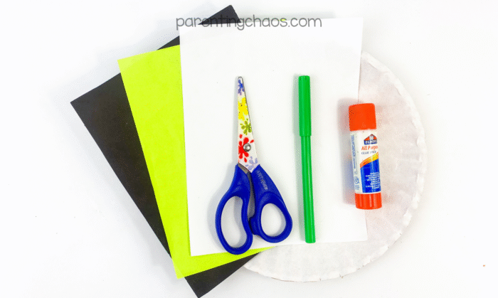 Supplies for the eyeball paper plate craft including black, green, and white construction paper, scissors, green marker, glue stick, and a white paper plate.