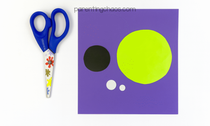 Green, black, and white paper circles cut out for the iris, pupil, and highlights of the eyeball craft, next to a pair of scissors.