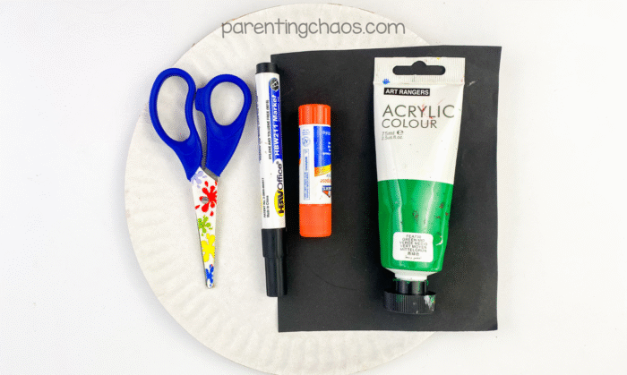 Paper plate craft supplies including scissors, glue stick, black marker, green acrylic paint, and black construction paper arranged on a paper plate.