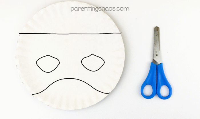 Paper plate with Frankenstein mask outline drawn in black marker, showing cut lines for eyes, mouth, and forehead, with scissors beside it.