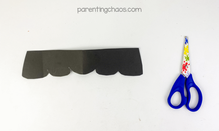 Black construction paper cut into a zig-zag hair shape with scissors beside it for Frankenstein mask craft.