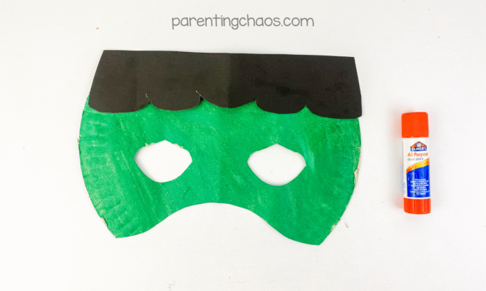 Frankenstein paper plate mask with black hair glued to the top, shown next to glue stick.