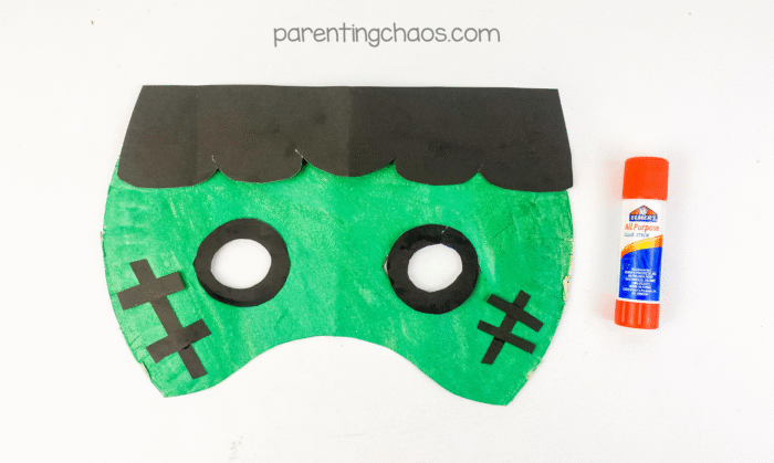Frankenstein paper plate mask with eyes and stitches glued on, next to a glue stick.