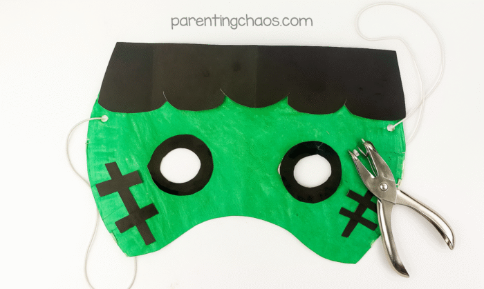 Frankenstein paper plate mask with holes punched on the sides and string threaded through, next to a hole punch.