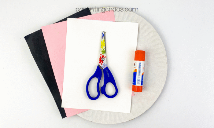 Craft supplies for ghost paper plate craft — black, pink, and white paper, scissors, glue stick, and paper plate.