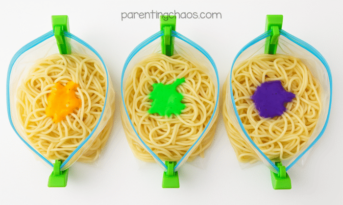 Three zipper bags filled with plain cooked spaghetti and bright splashes of orange, green, and purple dye on top, ready to be mixed for a Halloween sensory bin.