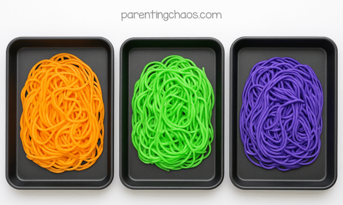 Three trays with dyed spaghetti in Halloween colors—orange, green, and purple—lined up side by side.