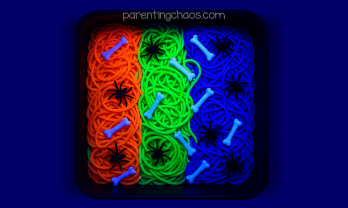 The same Halloween spaghetti sensory bin glowing under a black light, with neon orange, green, and blue spaghetti shining brightly.